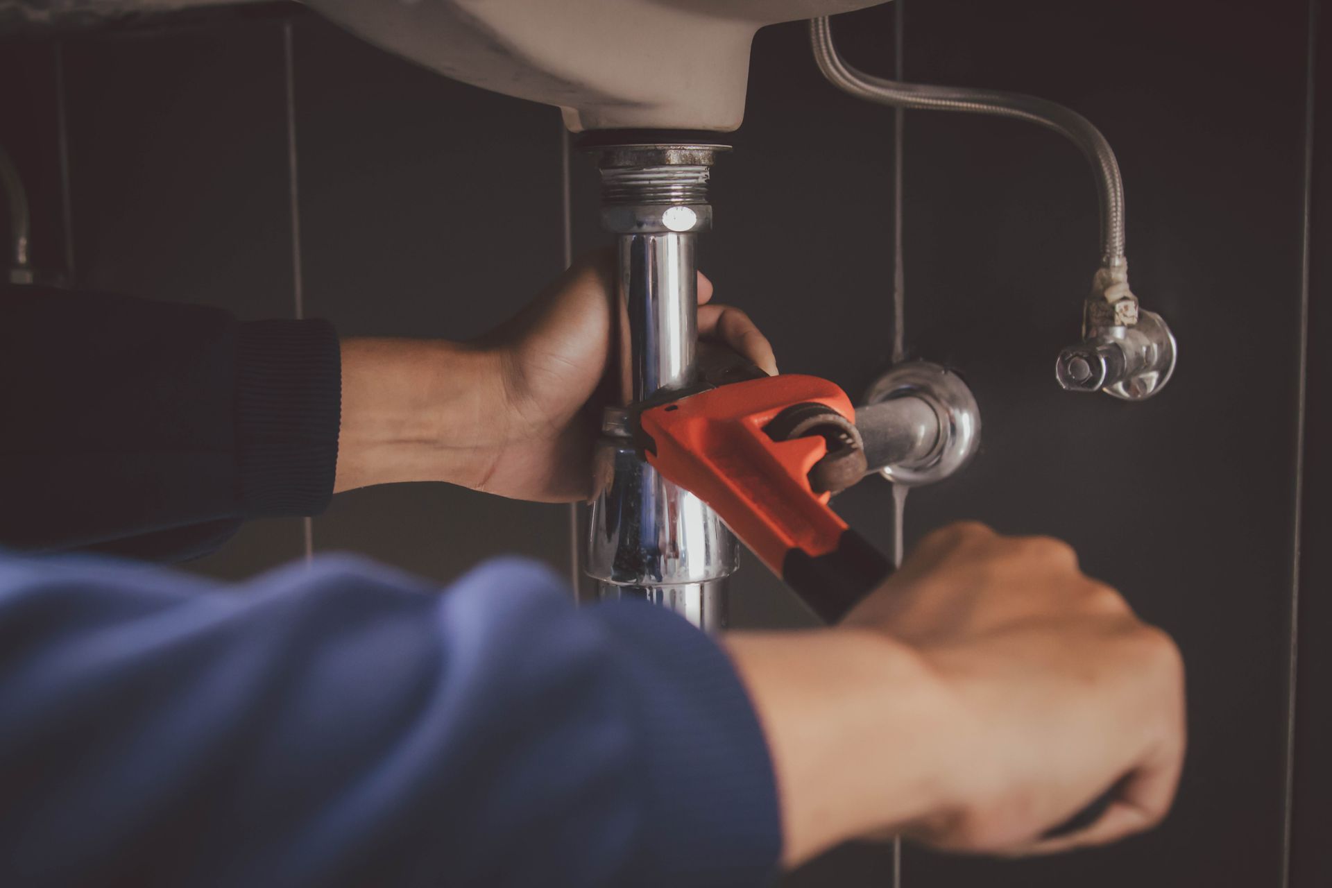 A person using a wrench to work on a sink's pipes in a dark bathroom.