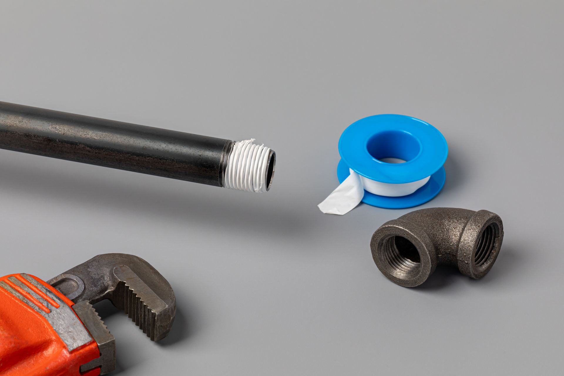 Plumbing tools on a gray surface: a wrench, black pipe with Teflon tape, a fitting, and a roll of tape.