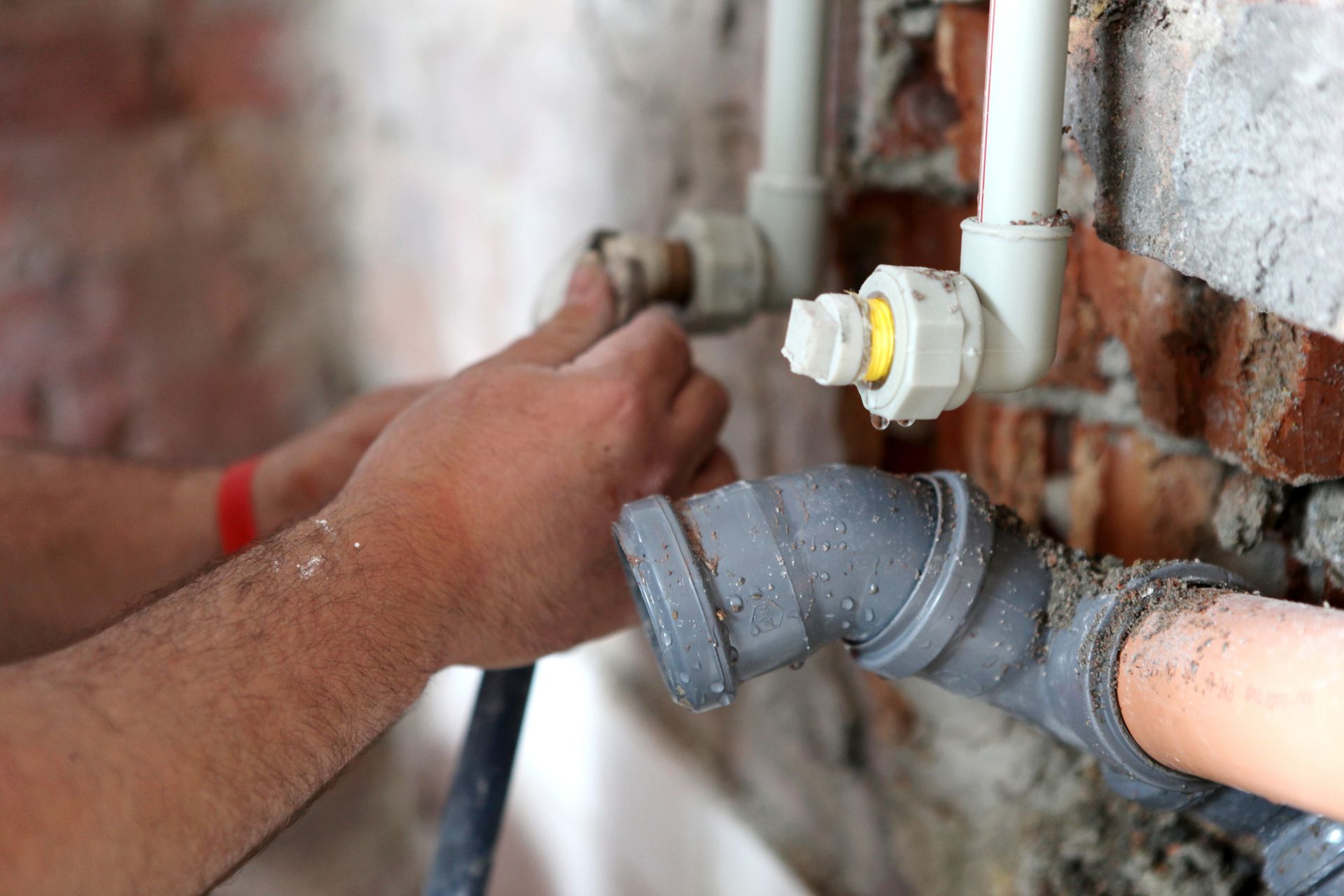 Hands of a plumber working on pipes near a brick wall. One hand holds a pipe, the other adjusts a fitting.