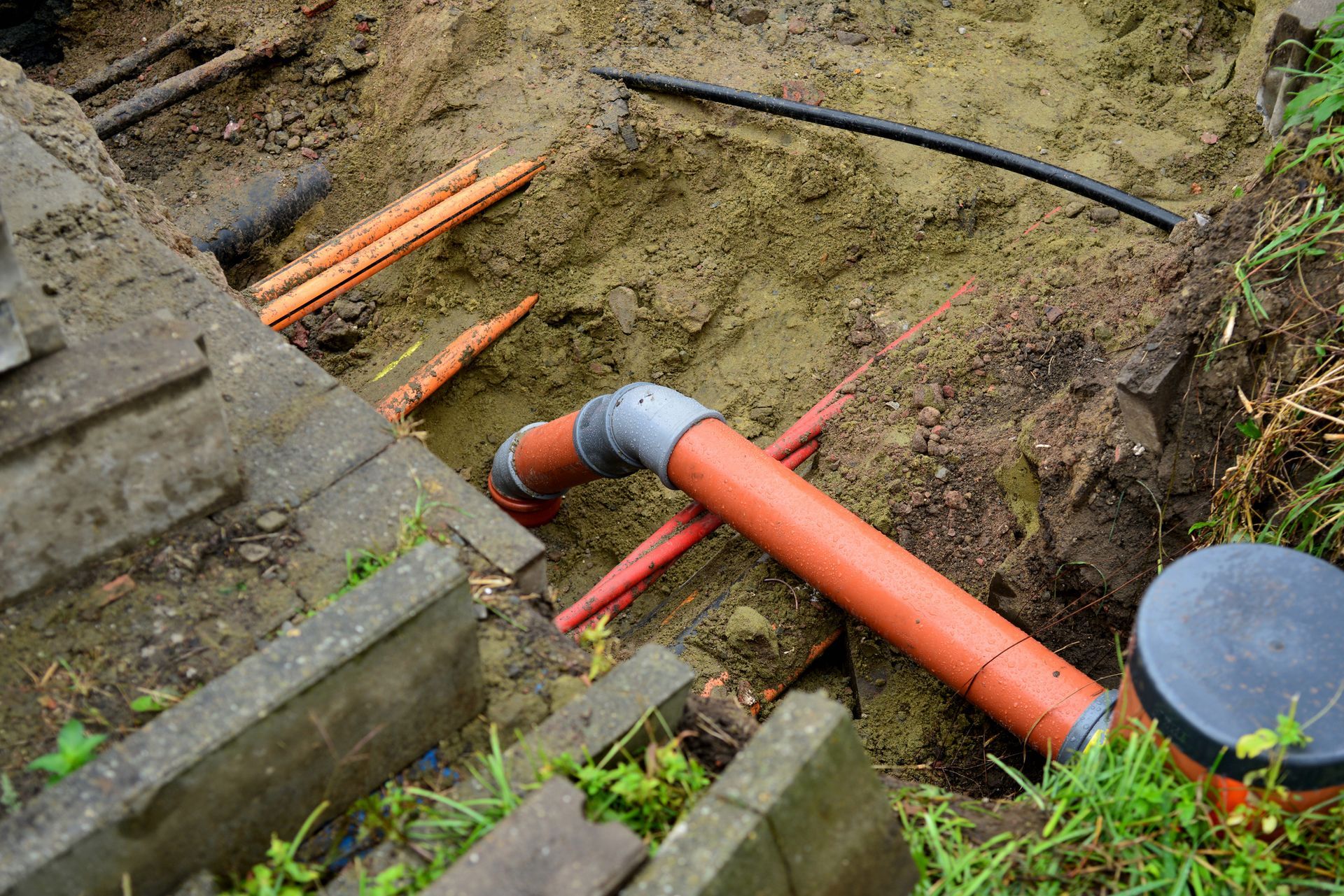 Orange and gray pipes laid in a dirt trench with other utility lines, next to a concrete structure.