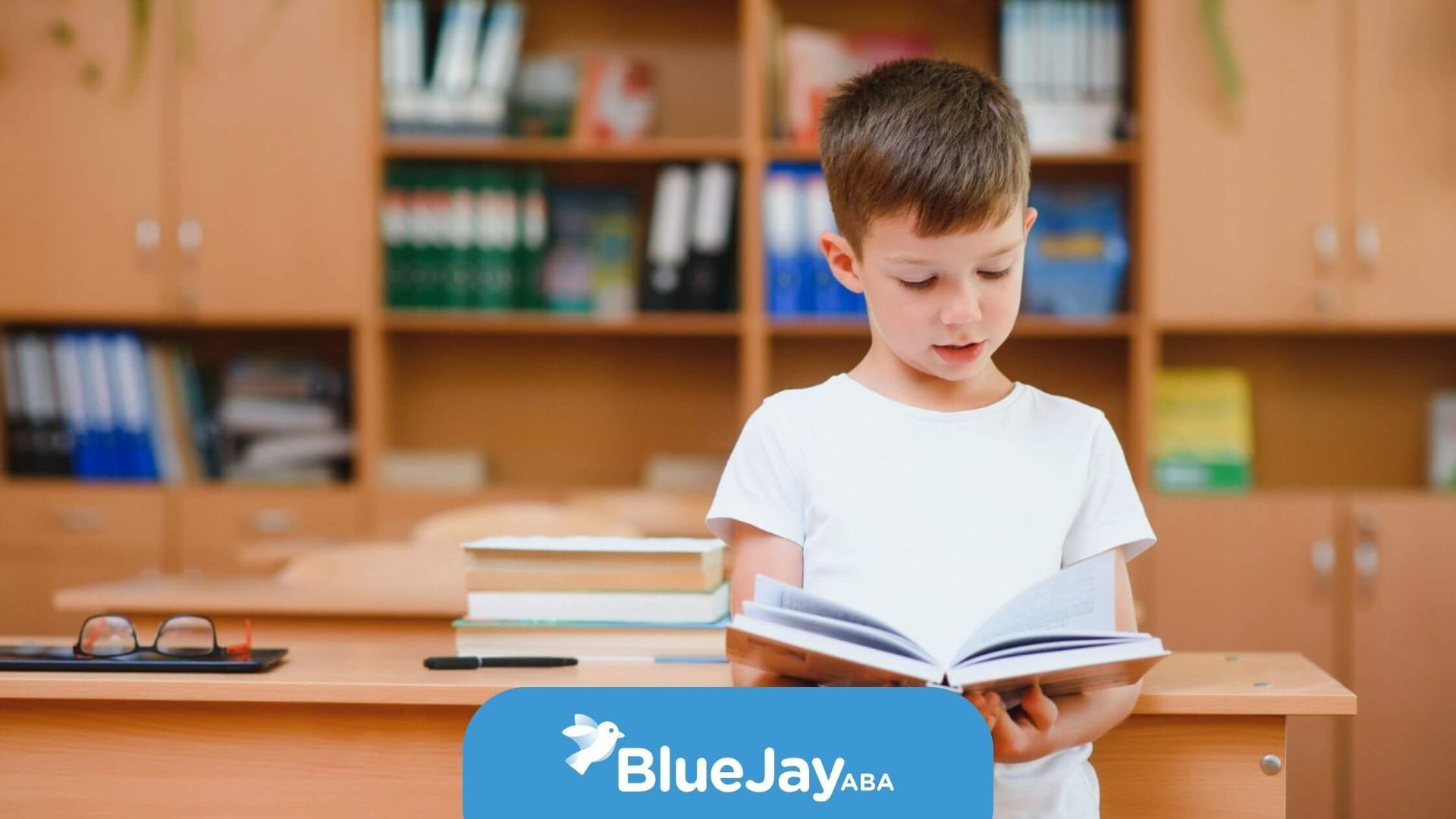 A young boy in a white shirt reading a book in a classroom before ABA therapy session.