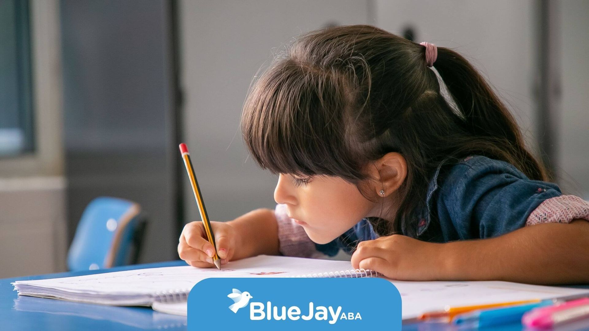 A young autistic girl sitting at a desk, concentrating as she draws with a red pencil during therapy