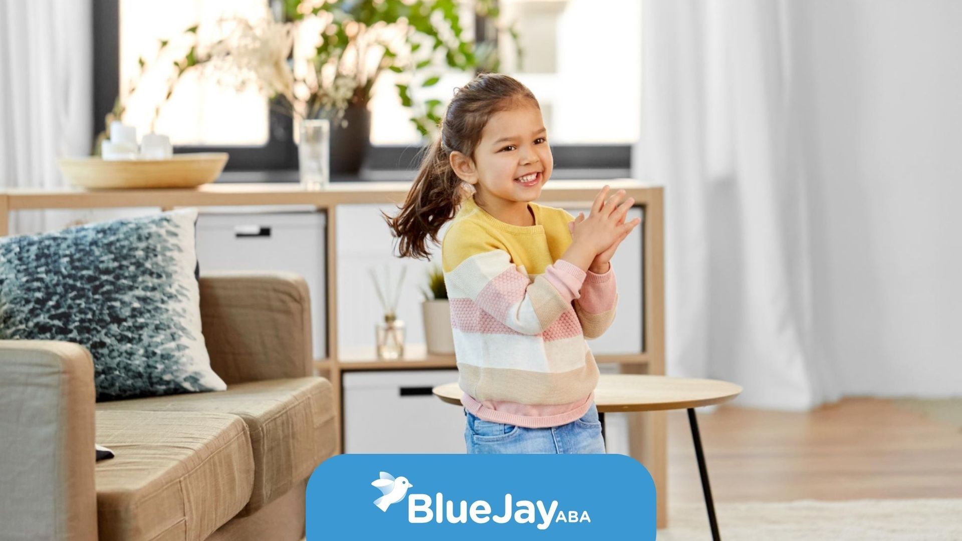 Child smiling and clapping in a bright living room with sofa, table, and flowers on a cabinet.
