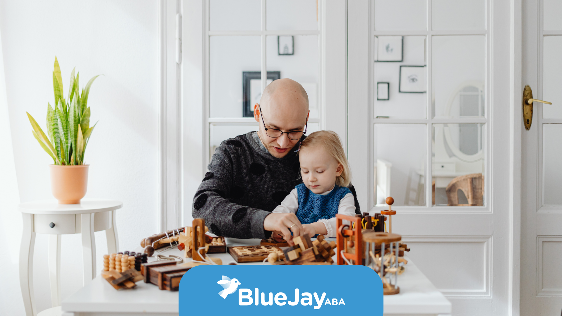 A person helps a young child play with wooden toys at a white table in a bright room.
