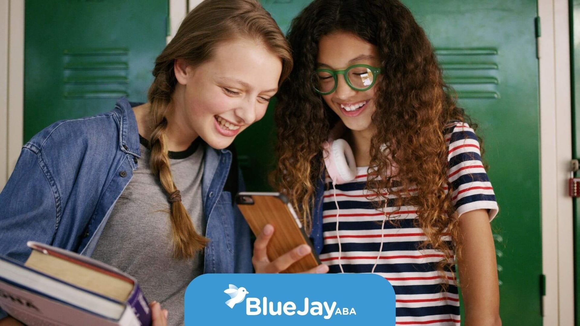 Two teenage girls with autism smiling and looking at a smartphone by school lockers.