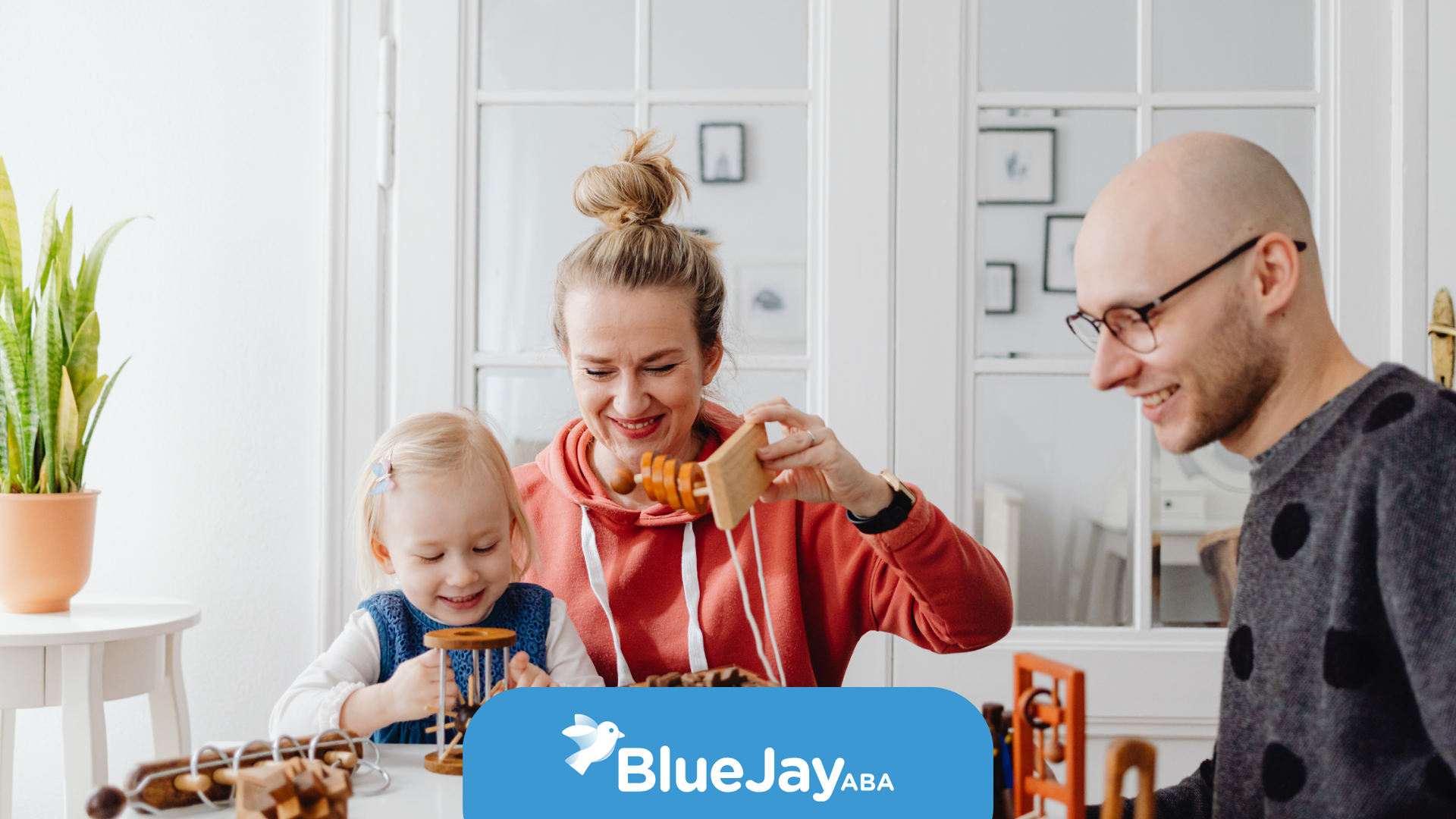 Family playing with wooden toys at a white table. The mother smiles while holding a toy, and the father wears glasses.