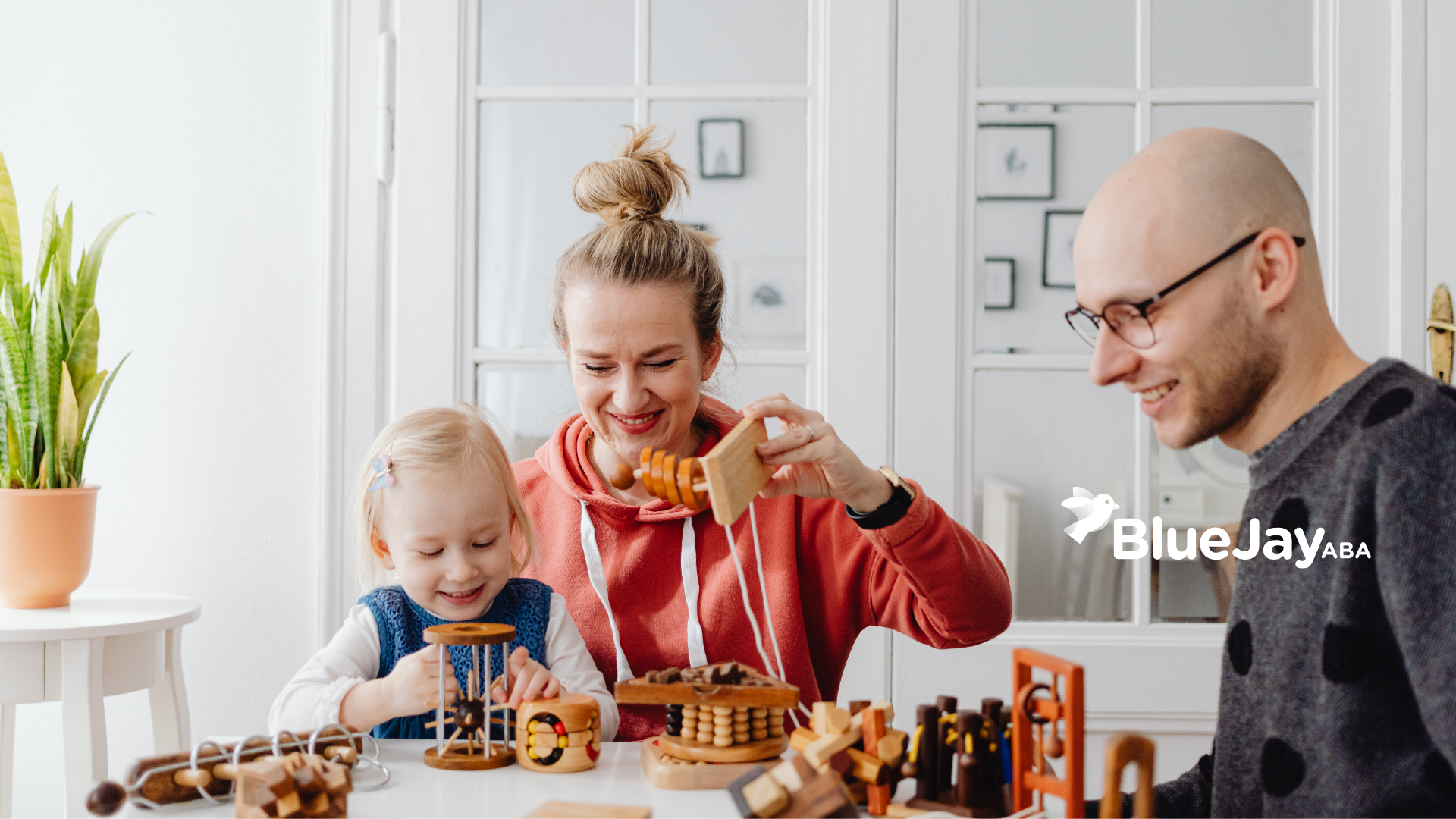 Family playing with wooden toys at a white table. The mother smiles while holding a toy, and the father wears glasses.