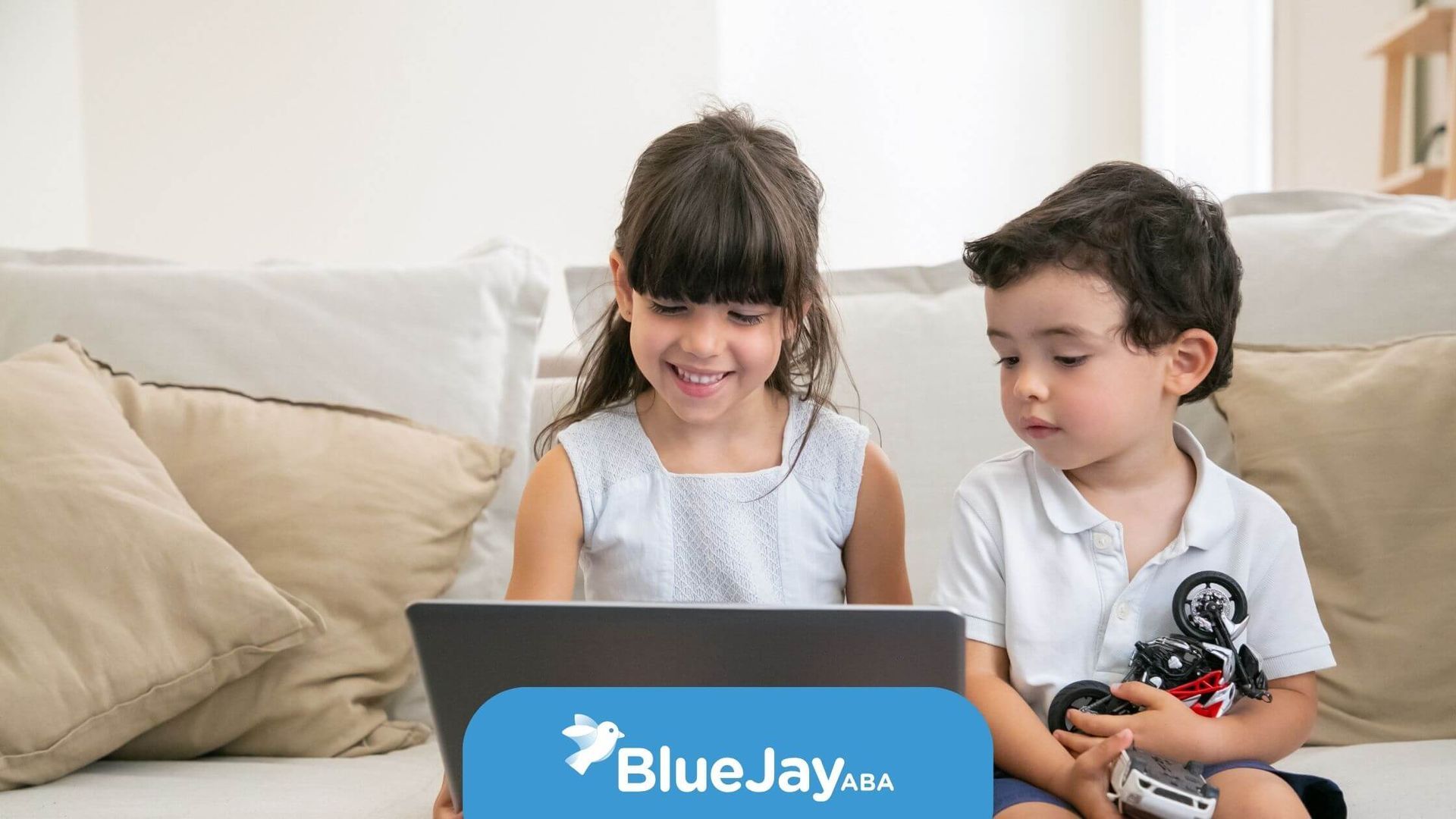 A girl & boy sitting on a couch, the girl is smiling while using a laptop, & the boy is watching.