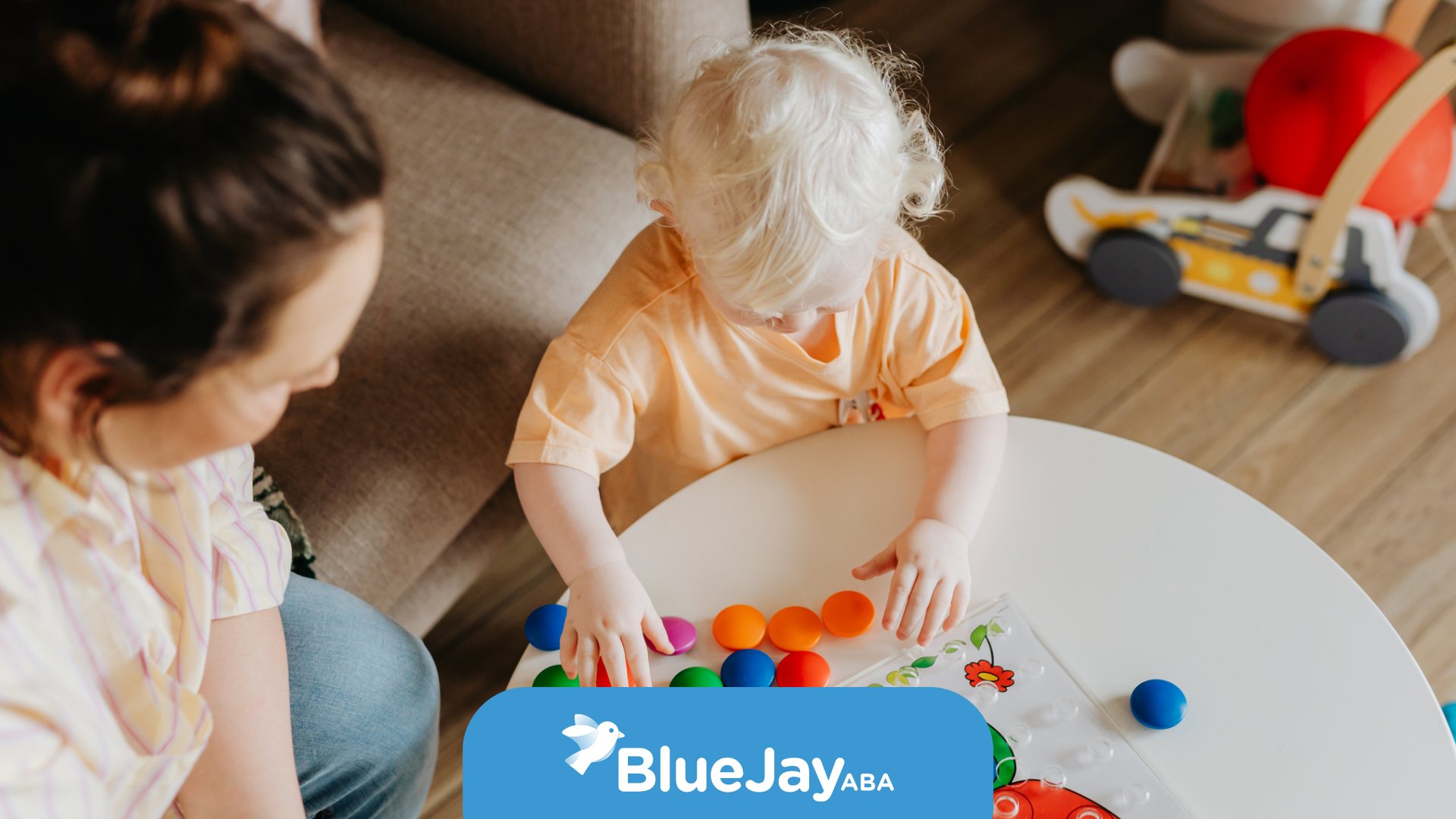 Woman watches toddler playing with colorful balls on a white table indoors.