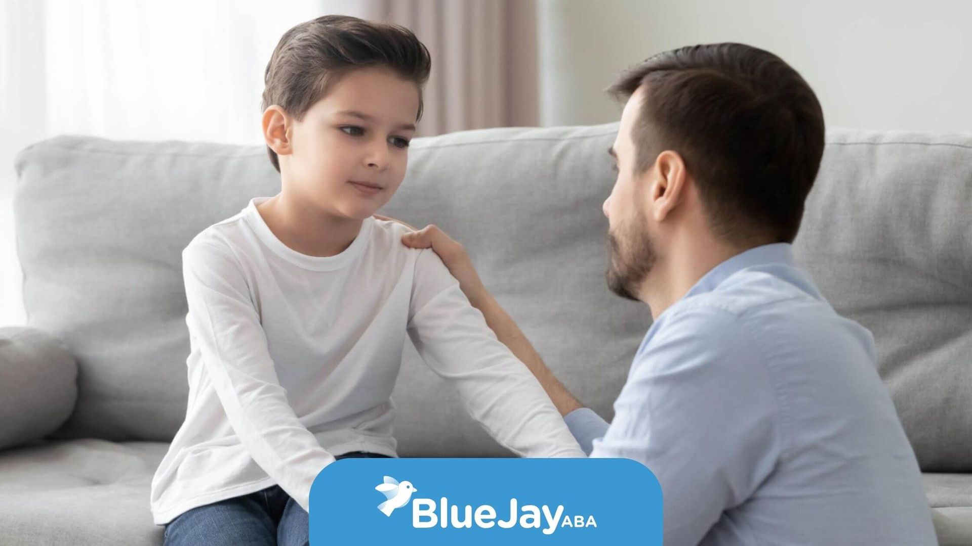 BCBA gently holding autistic son's arms while talking on a couch in ABA therapy.