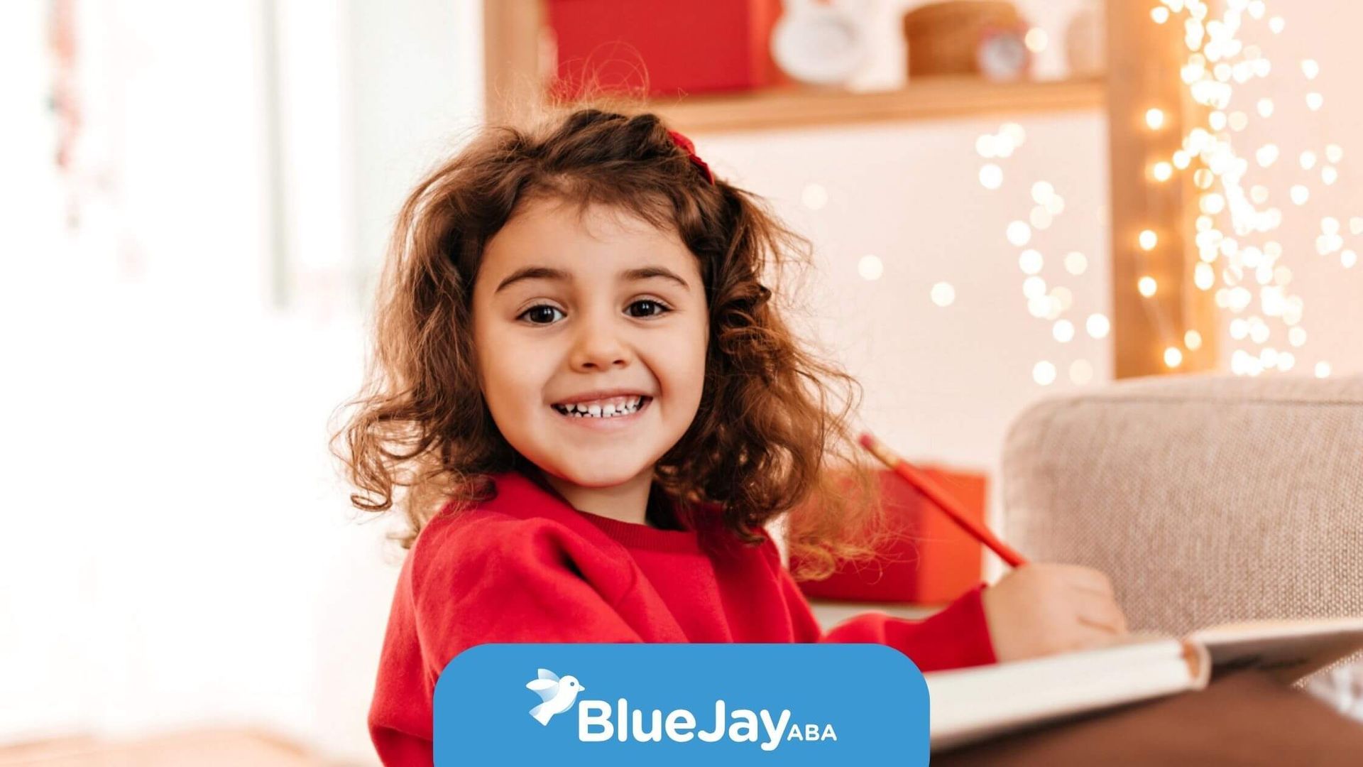 A smiling young girl with curly hair wearing a red sweater and writing in a book during ABA therapy.