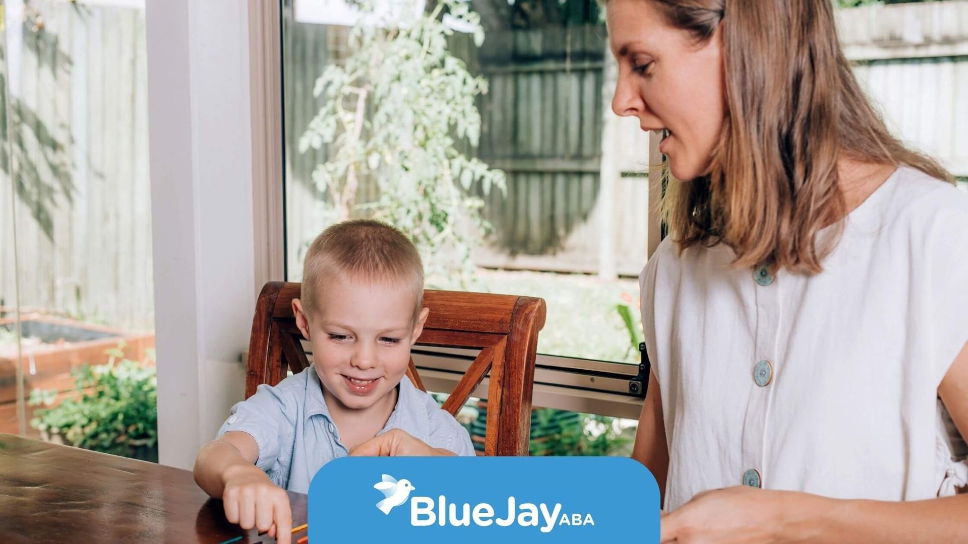 A boy & a woman sitting at a table, the boy is playing with colorful sticks, while the woman helps.