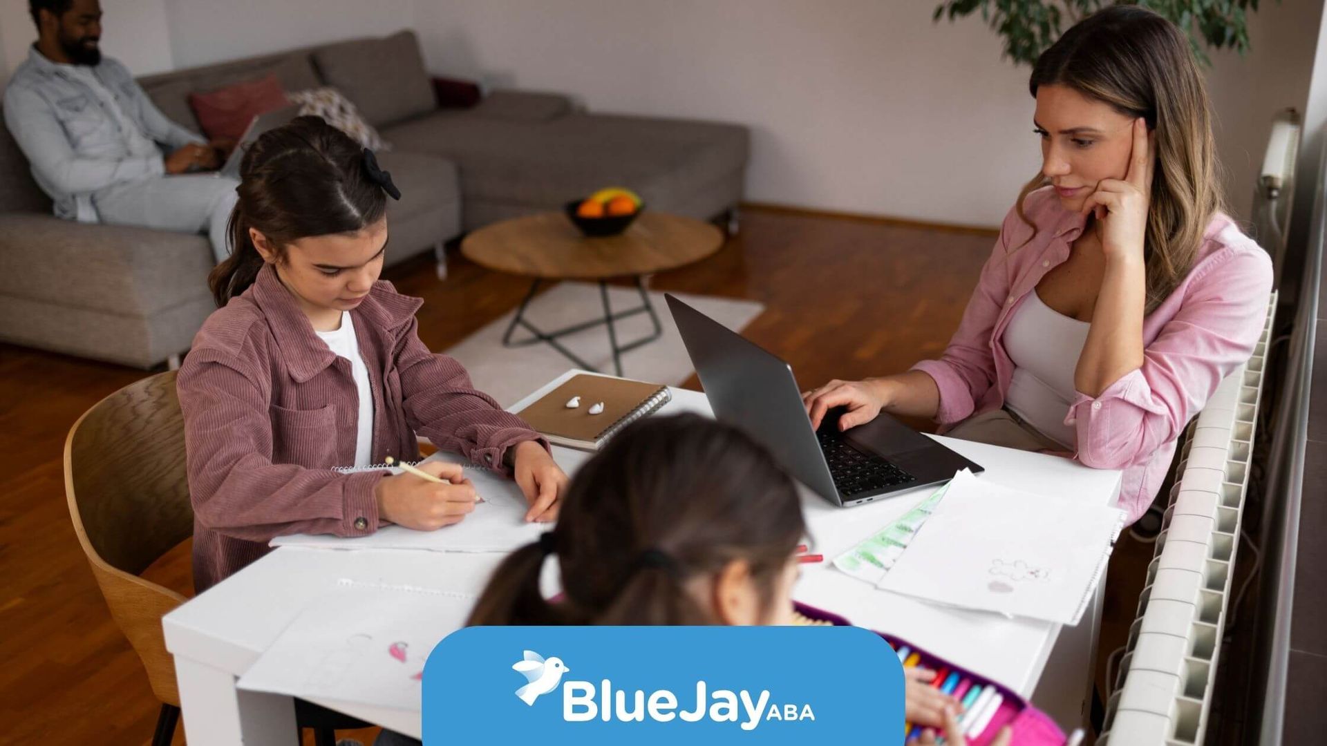 Family sitting at a table doing creative activities together, with a parent working on a laptop