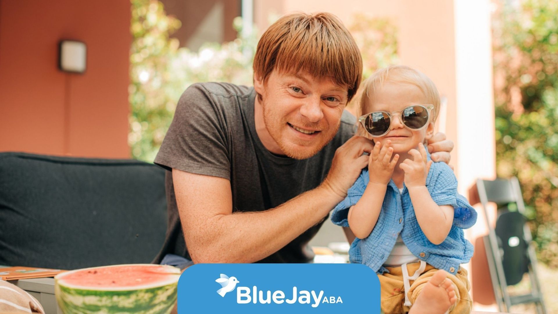 Man smiling with child in sunglasses at an outdoor patio table with watermelon slices and bowls