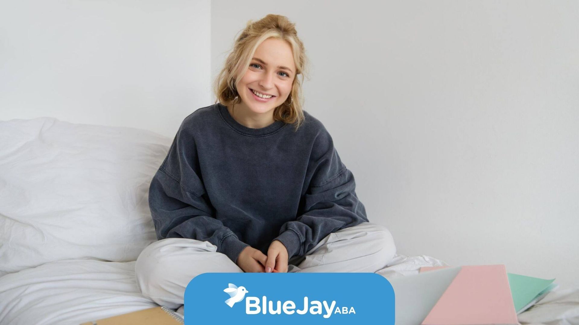 A smiling blonde woman with autism sits cross-legged on a bed with notebooks and a laptop.