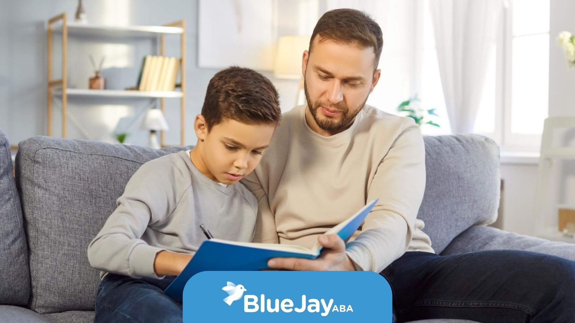 A male BCBA and a boy with autism sit on a grey sofa together in ABA therapy.