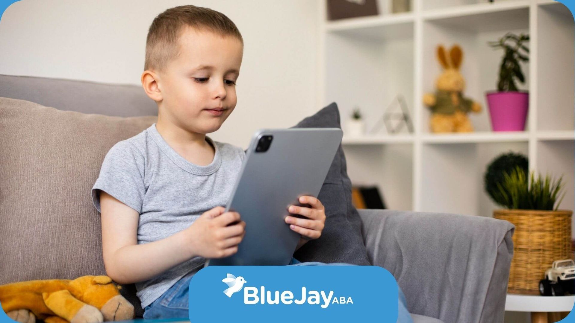 Young boy with autism sitting on a couch using an iPad with a toy and book beside him.