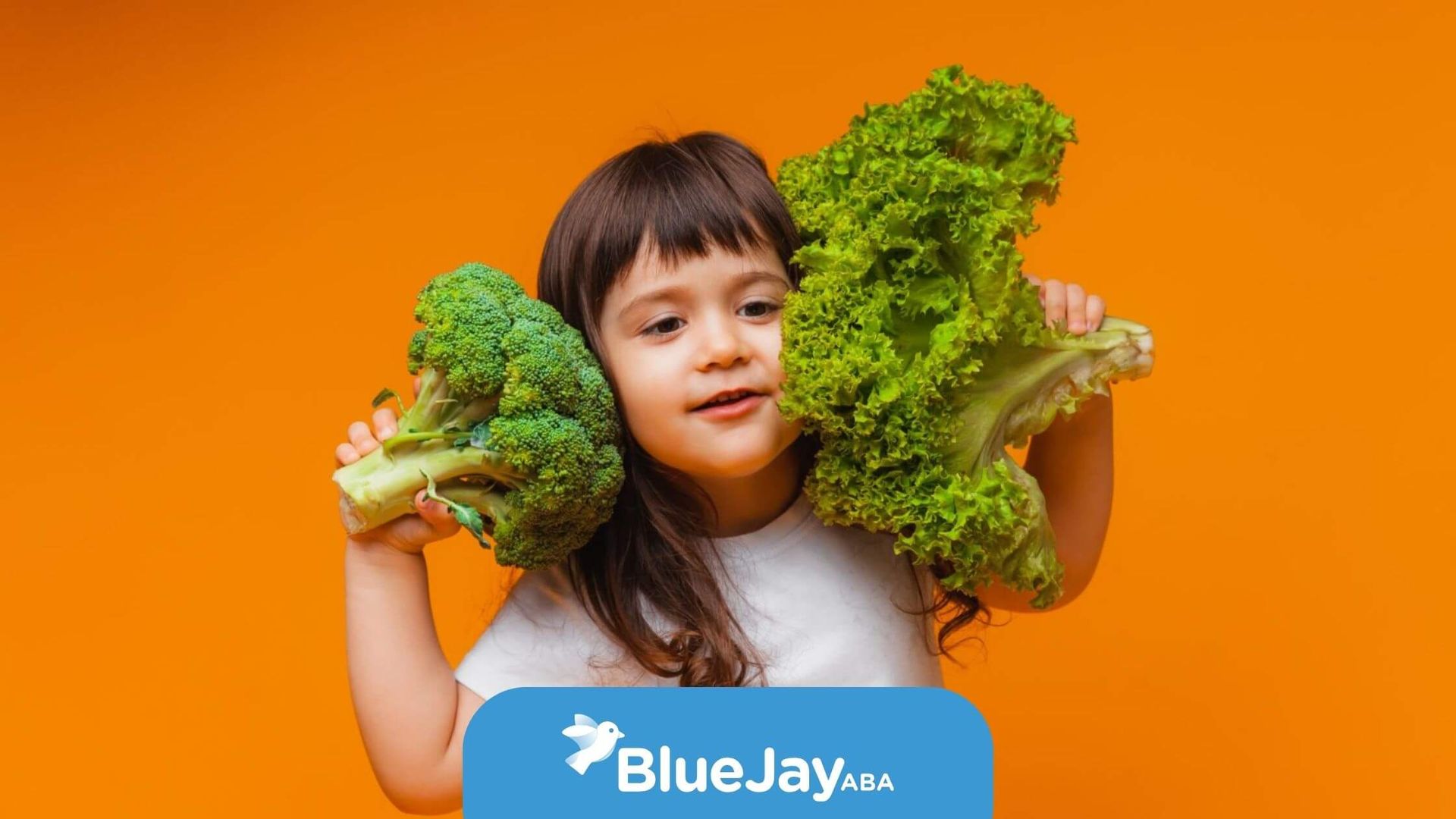 Child with autism holding broccoli and lettuce against orange background.
