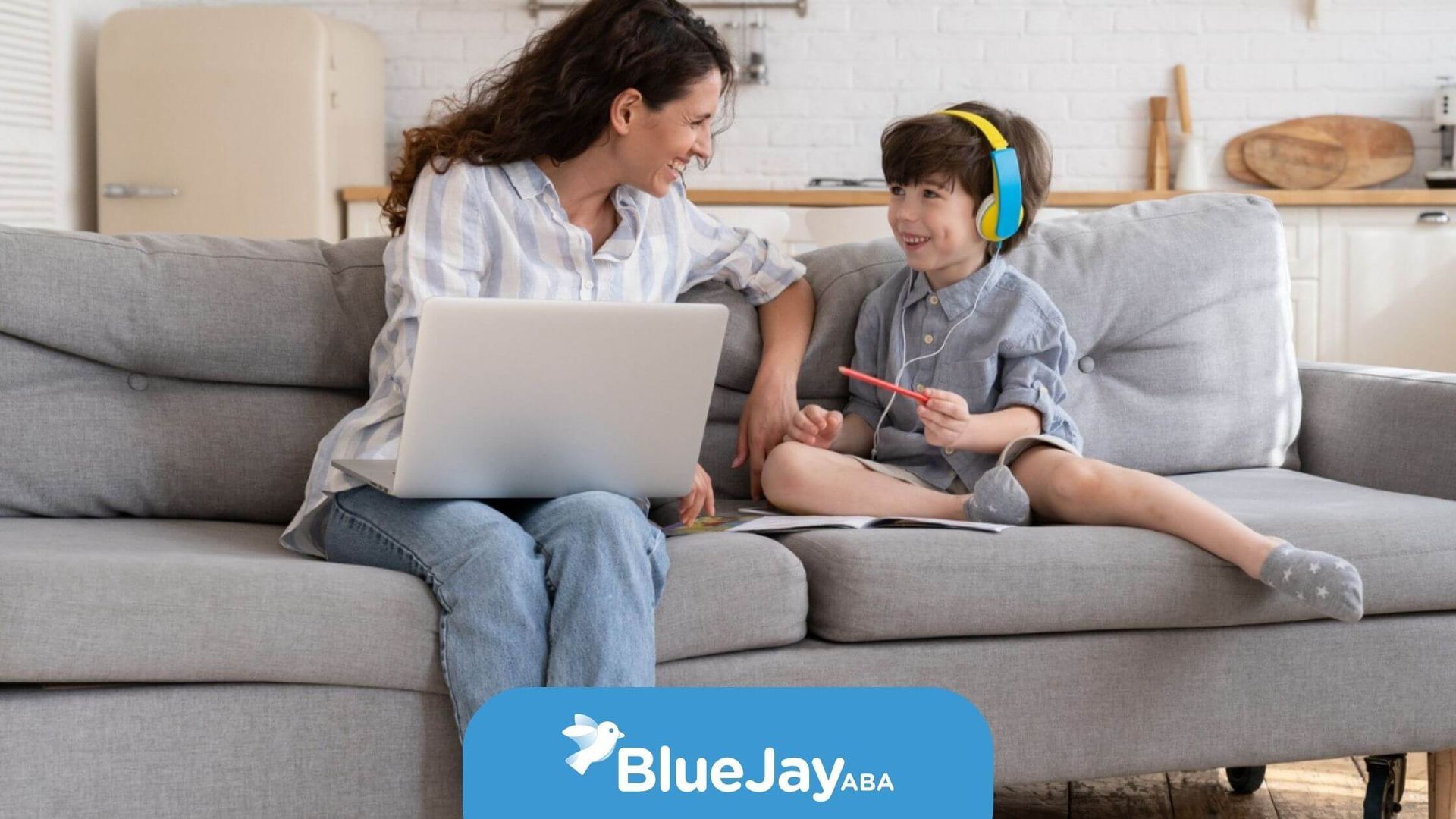 BCBA with laptop smiling at a child wearing headphones on a couch.