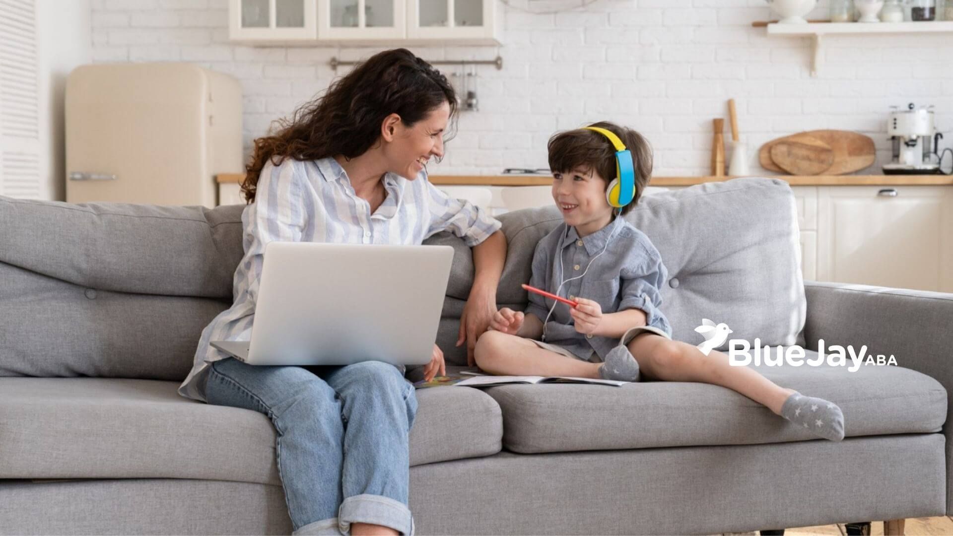 BCBA with laptop smiling at a child wearing headphones on a couch.
