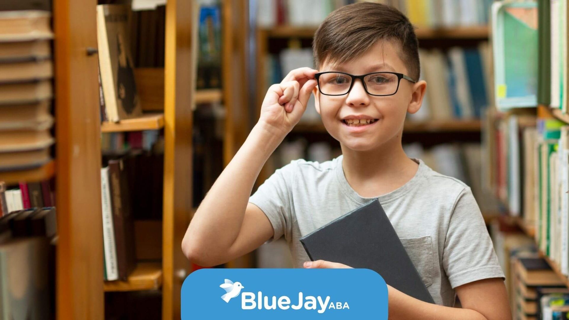 Smiling boy with glasses holding a book in a library.
