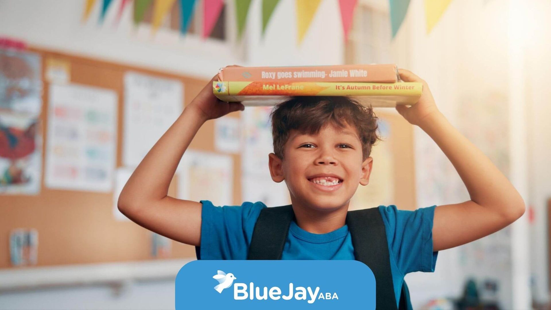 Boy with books balanced on head, smiling. Wearing backpack in classroom with colorful decorations.