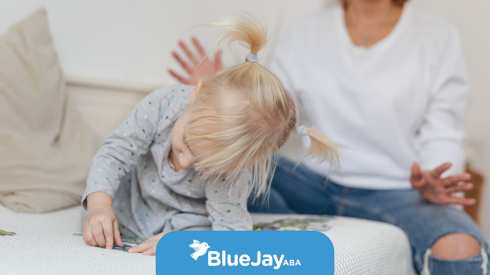 Blonde toddler bends down on a bed; adult reaches out.