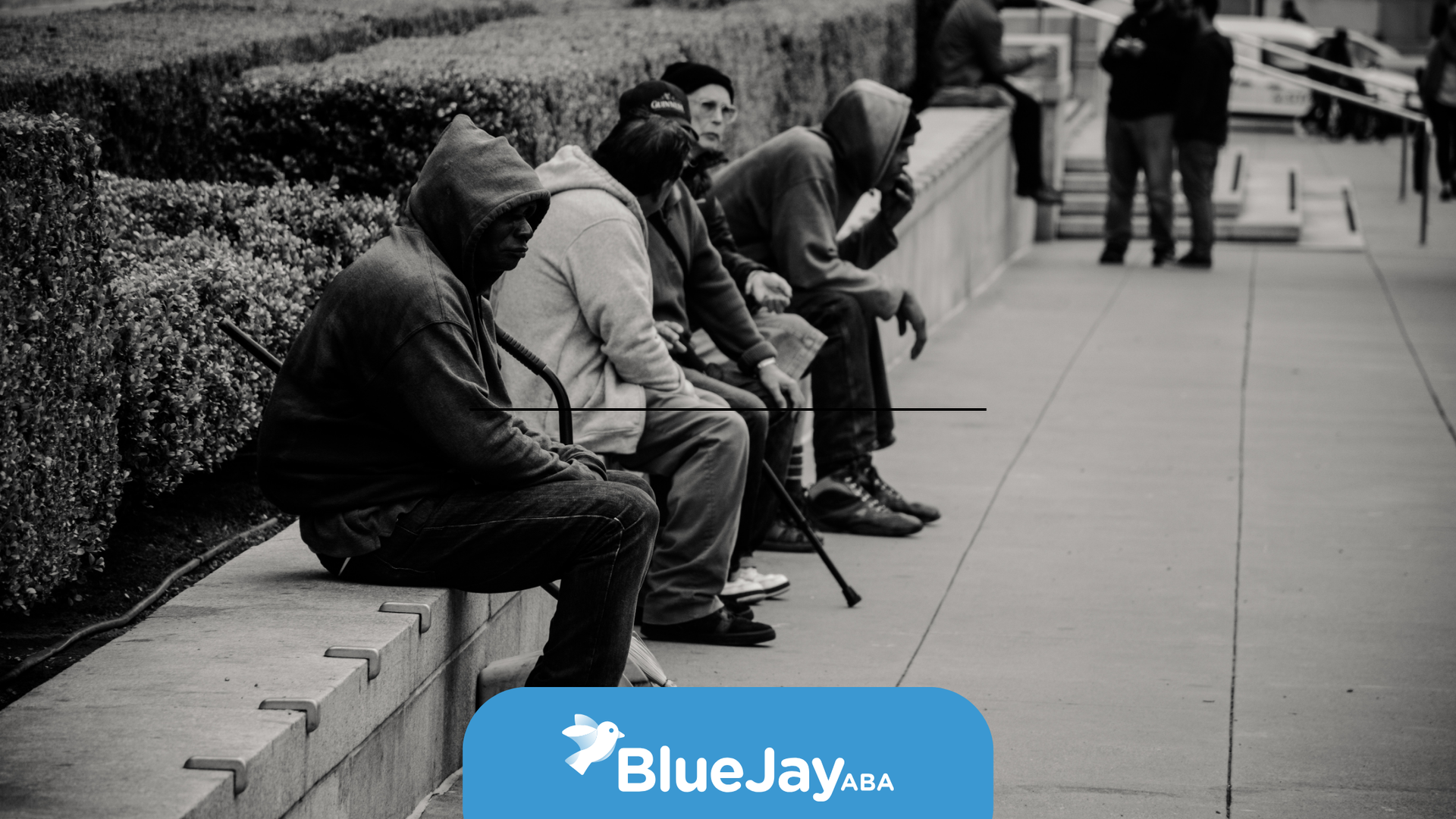 Five people in hoodies sit on a concrete ledge. One wears a mask. A blurred couple stands in the background.