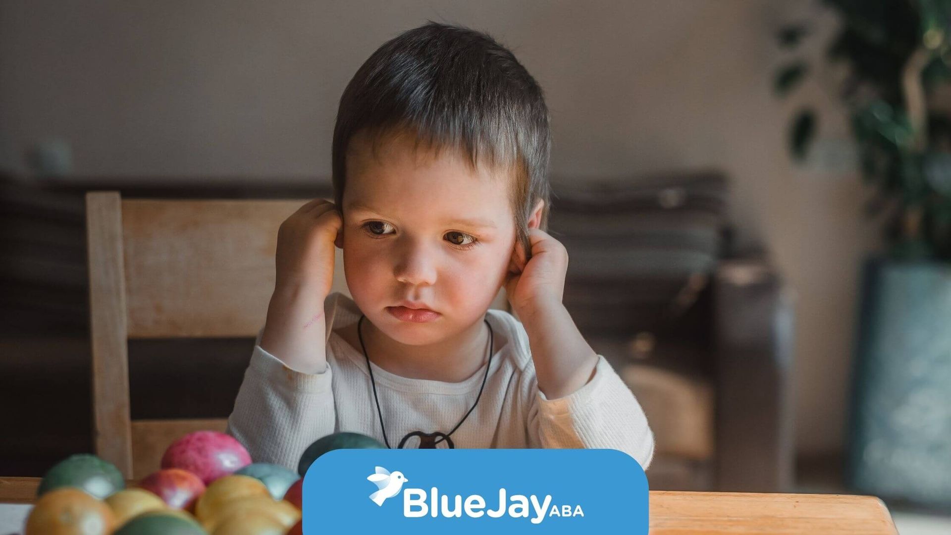 Autistic child holding his ears with a distressed expression, sitting near colorful Easter eggs.