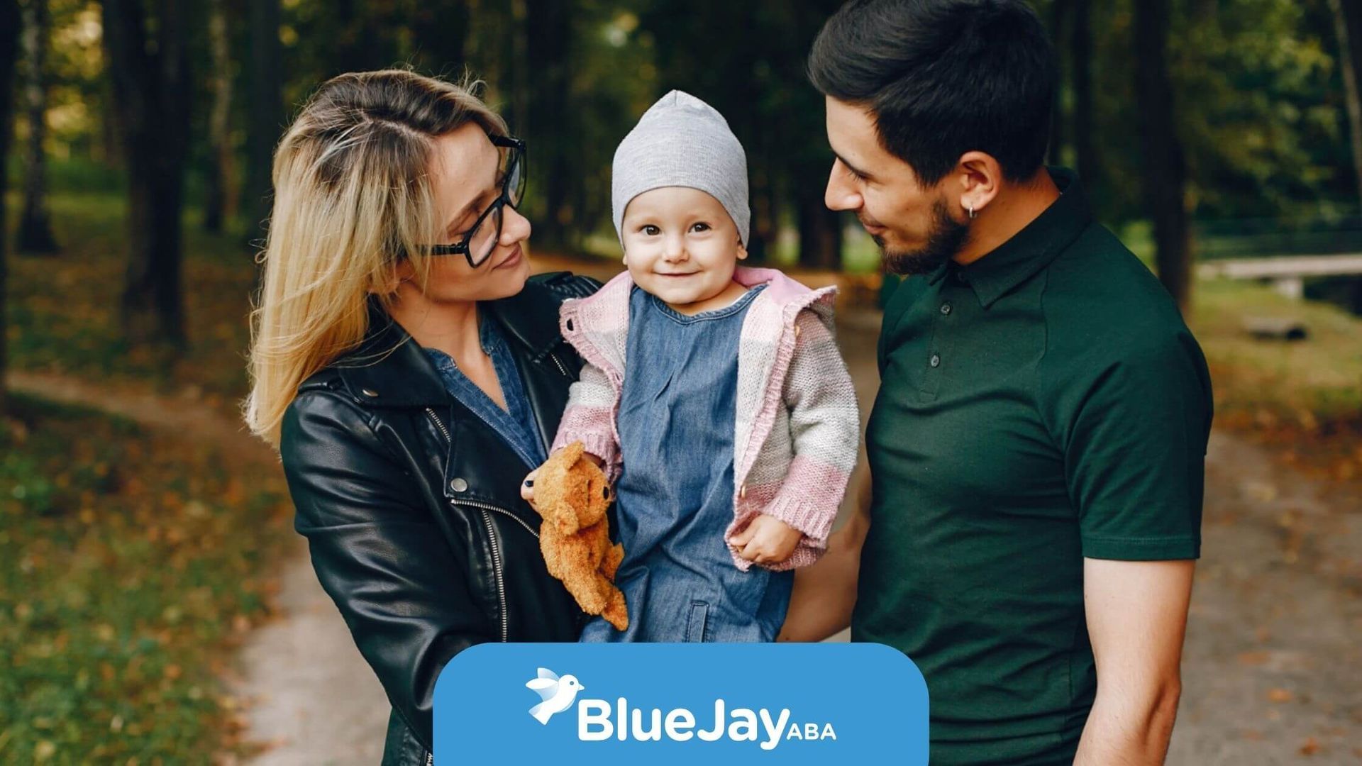 A mother and father smiling at their baby with autism holding a teddy bear outdoors.