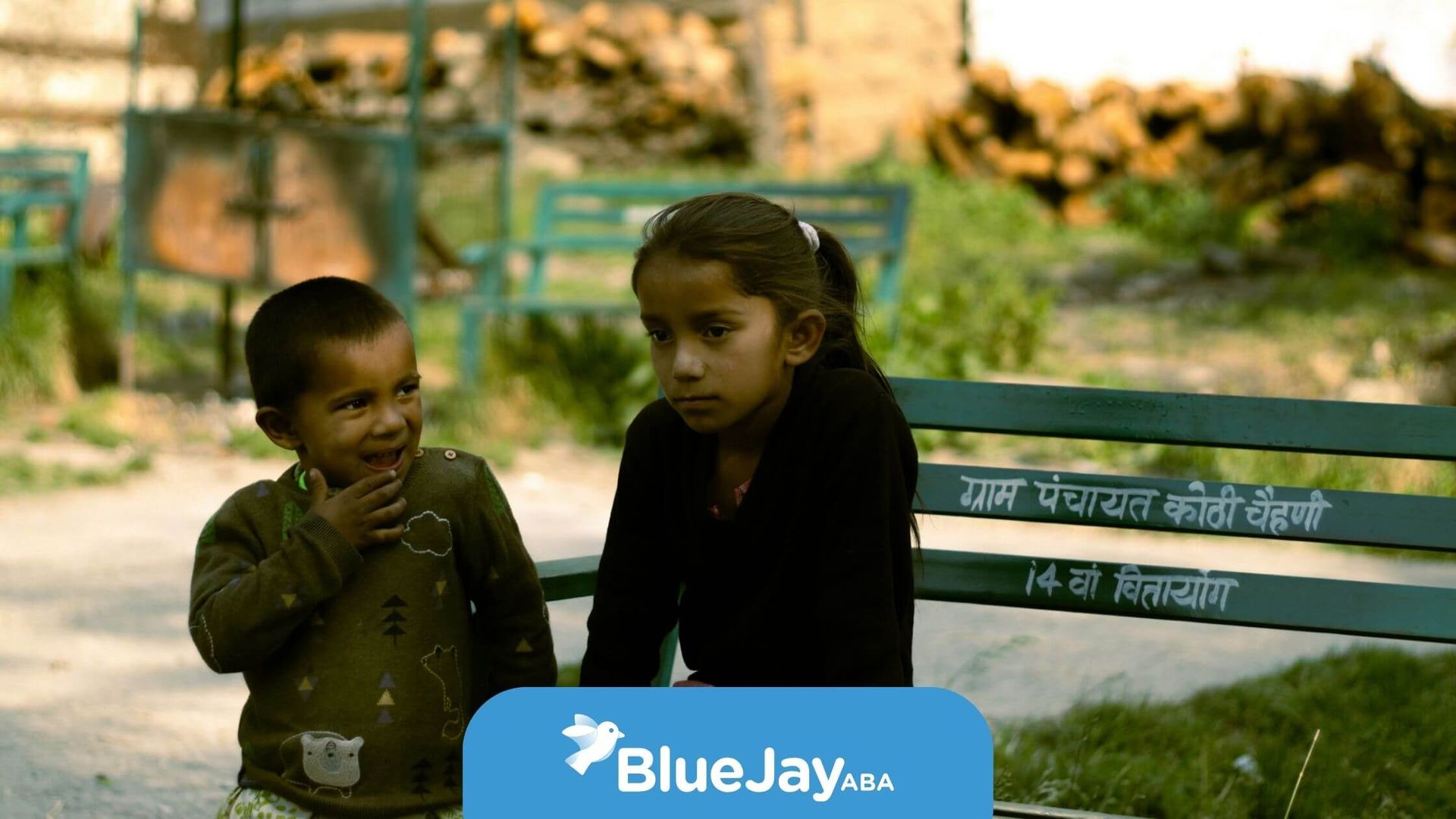 Two autistic children sitting on a green bench outdoors, one smiling and one looking serious.