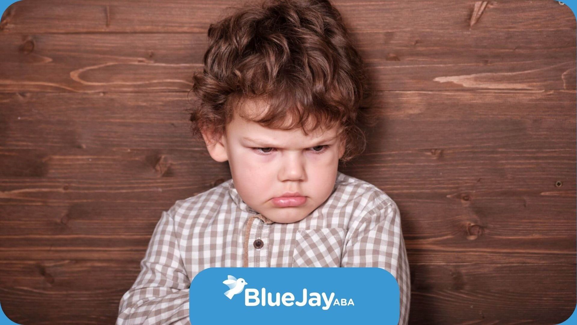 A young boy with autism is standing in front of a wooden wall with his arms crossed.