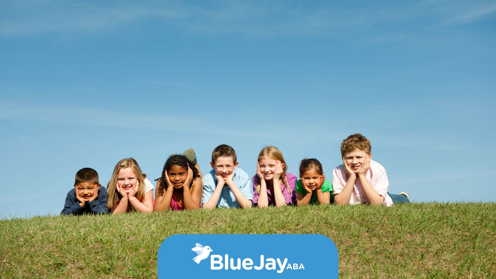 Children lying on a grassy hill, smiling at the camera under a blue sky.