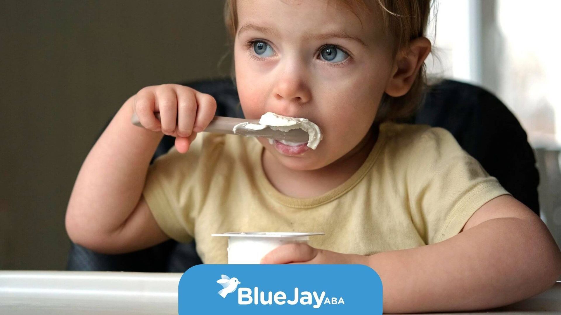 A young child with autism sitting in a high chair, using a spoon to eat yogurt.
