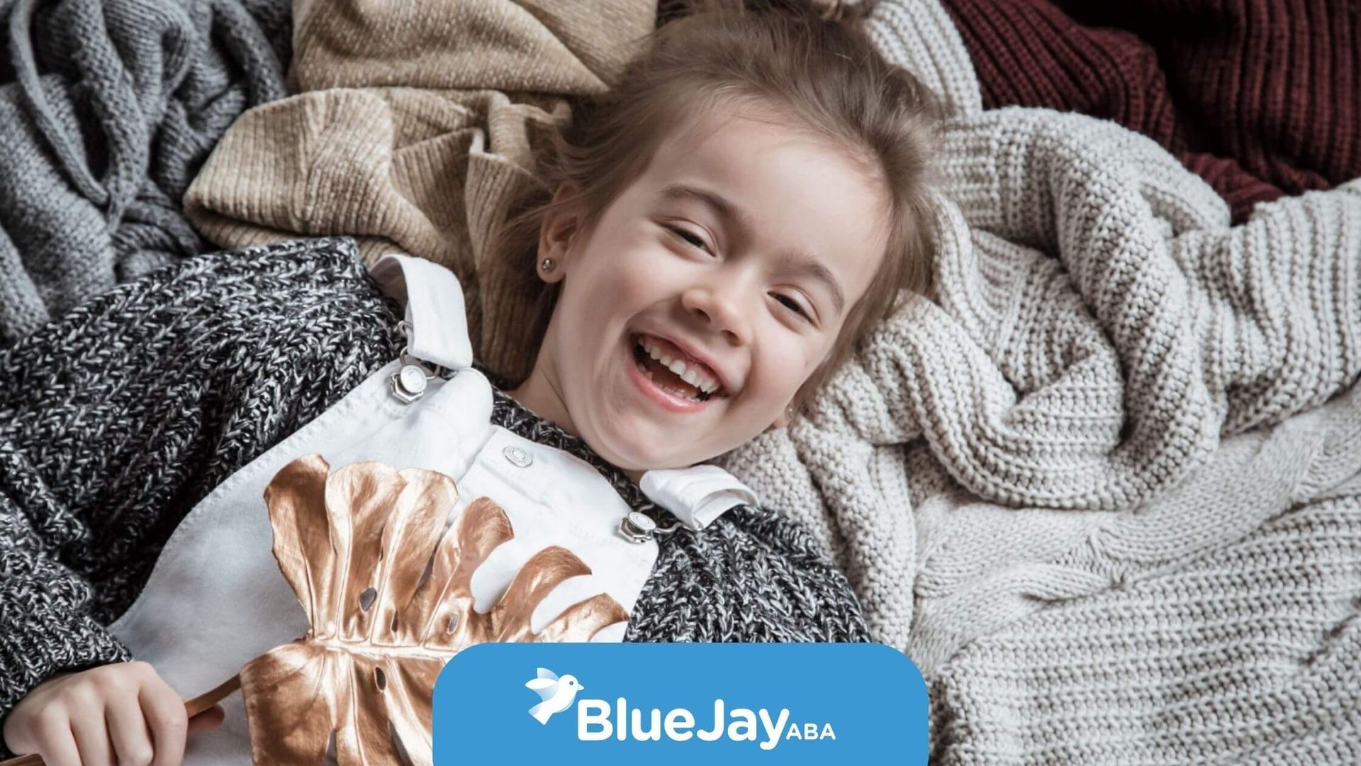 A laughing young girl lying down on a pile of various knit sweaters.
