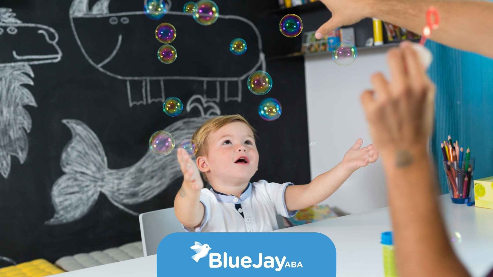 A young child reaches for floating bubbles in a room with a chalkboard drawing; happy expression.