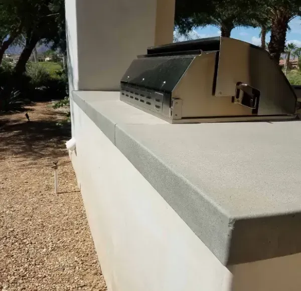 Outdoor countertop with built-in grill, light-colored walls, and gravel ground.