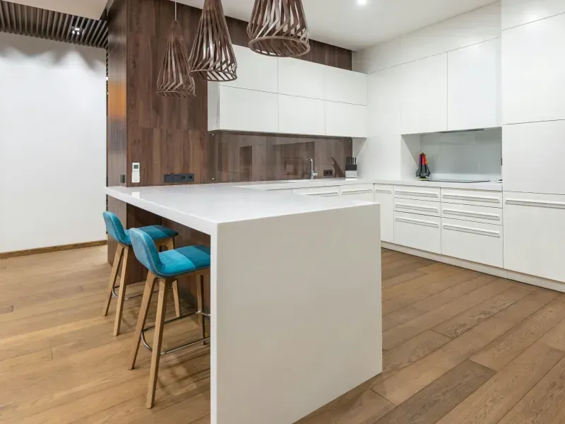 Modern white kitchen with a white island, wooden floors, blue bar stools, and wooden pendant lights.