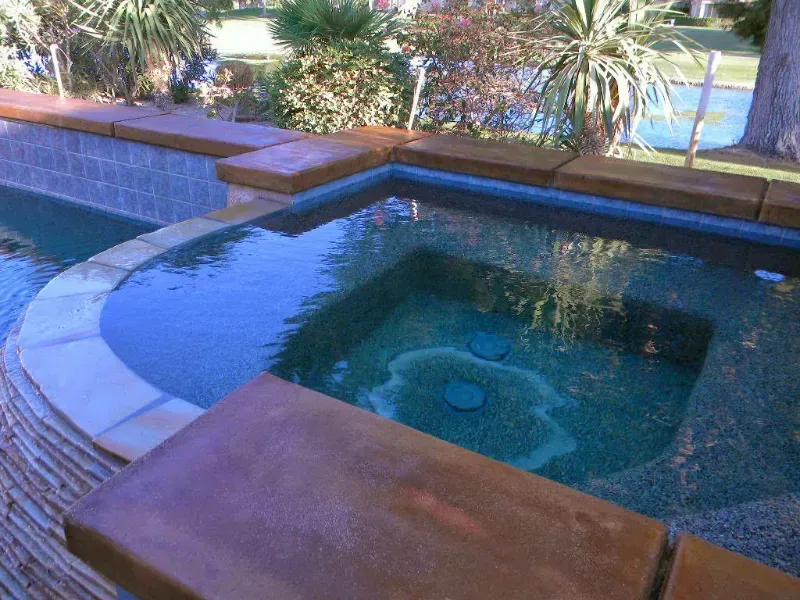 A rectangular spa pool with dark blue water and tan stone coping, set against a backdrop of garden greenery.