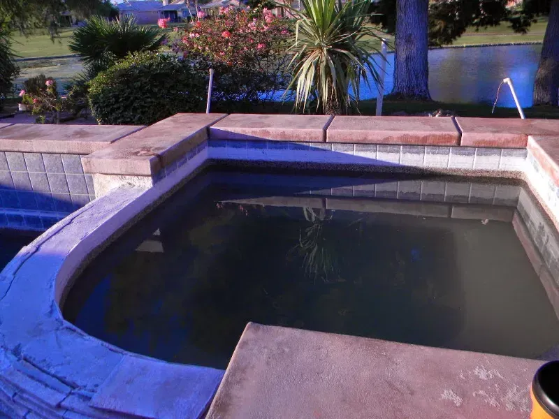 A square, tiled hot tub with murky water, surrounded by tan concrete coping and a landscaped backyard.