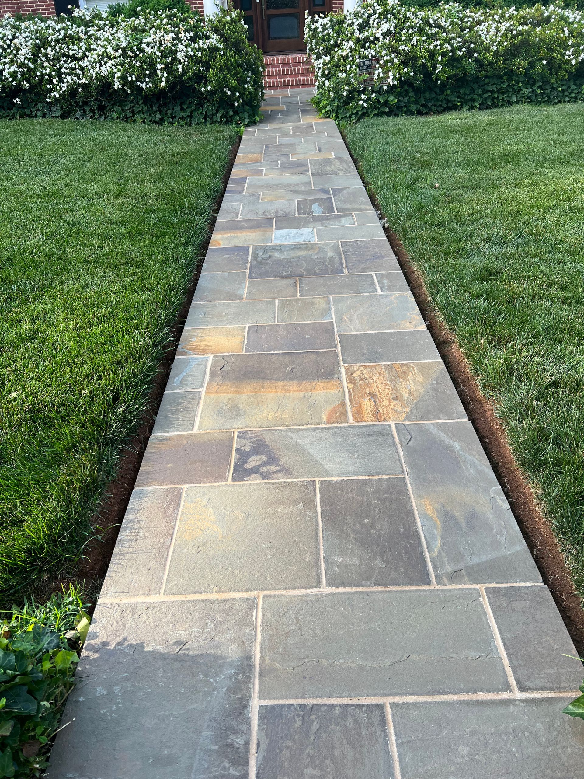 A stone walkway leading to a house surrounded by grass and bushes.