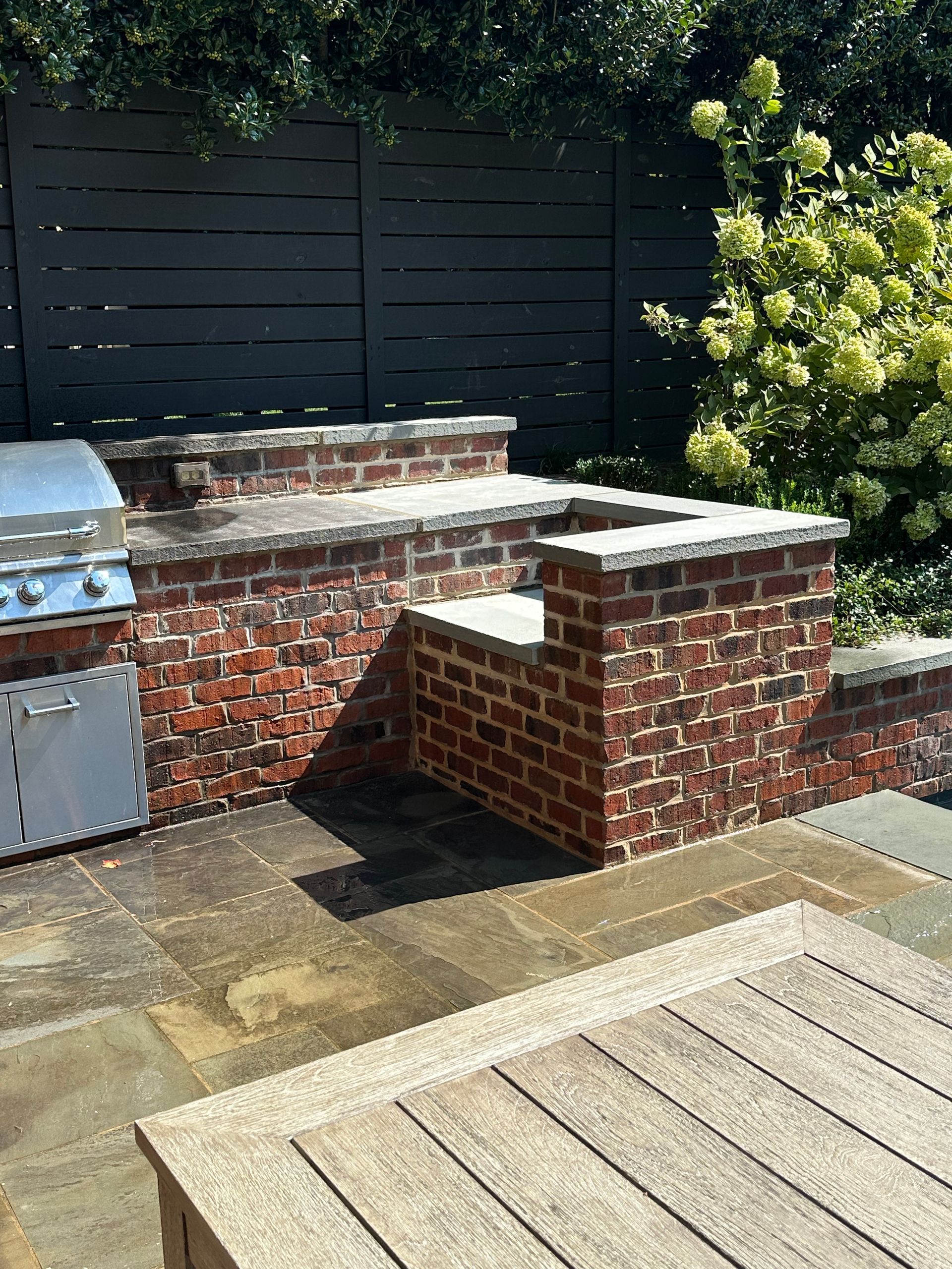 A brick wall with a grill and a wooden table in a backyard.