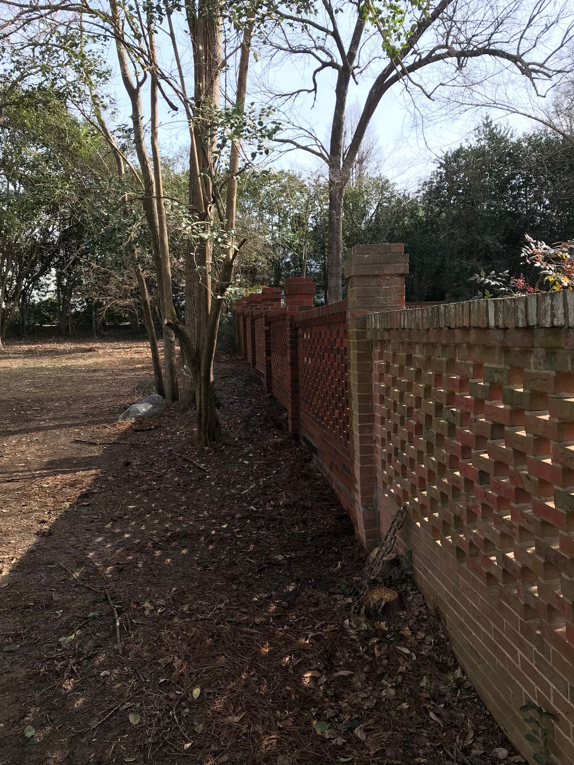 A brick fence with trees in the background and leaves on the ground.