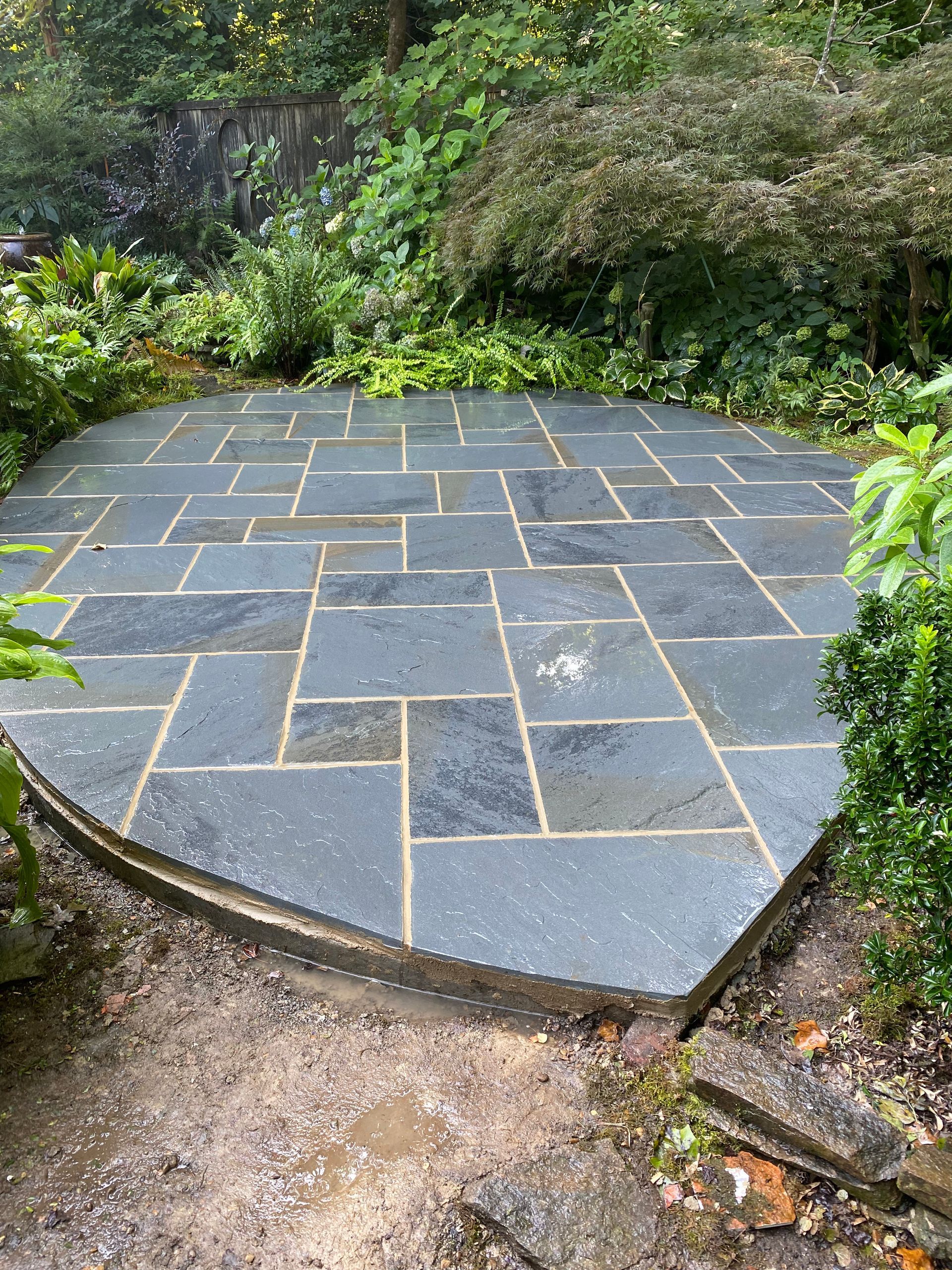 A circular stone patio in the middle of a garden surrounded by trees.