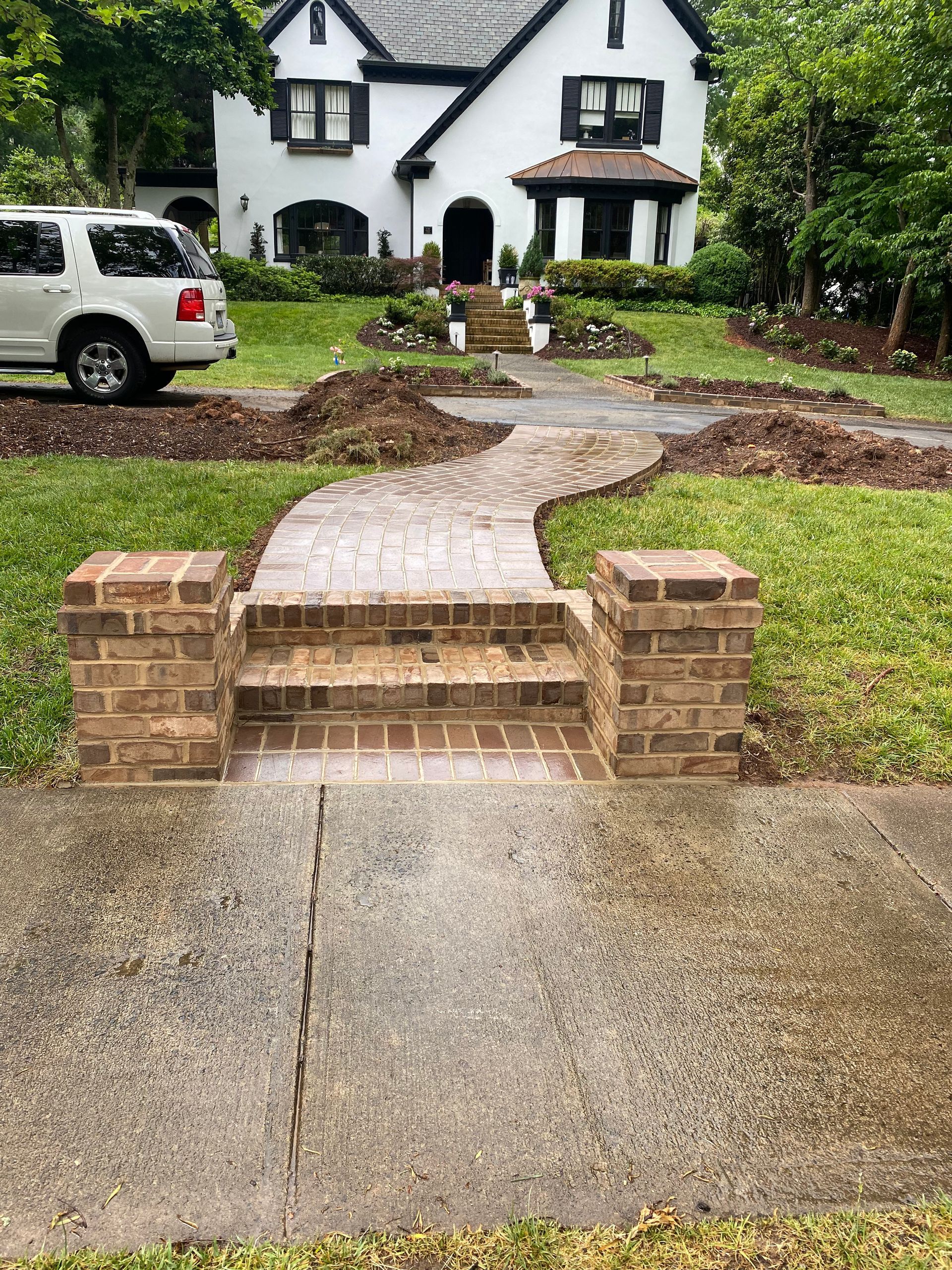 A white suv is parked in front of a white house with a brick walkway leading to it.