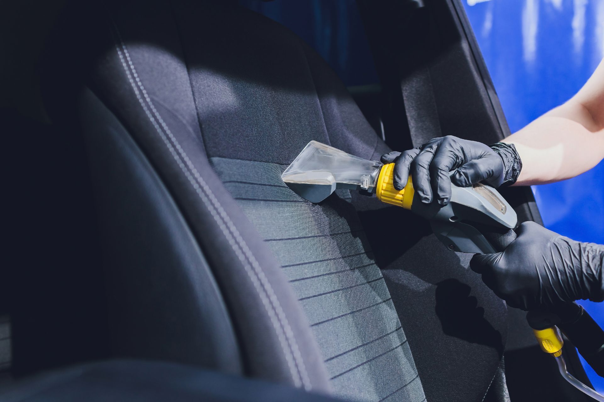 A person wearing black gloves uses a machine to deep clean a dark fabric car seat.