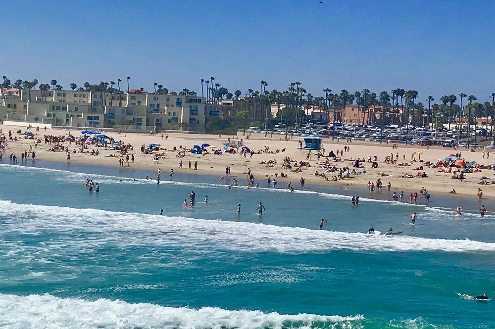 People on a sunny beach with buildings and palm trees. Turquoise waves crash onshore.