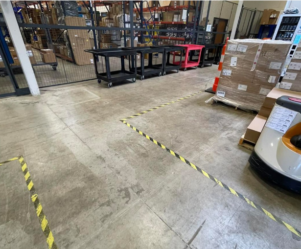 Warehouse floor marked with yellow and black hazard tape, showing work areas and pallets.