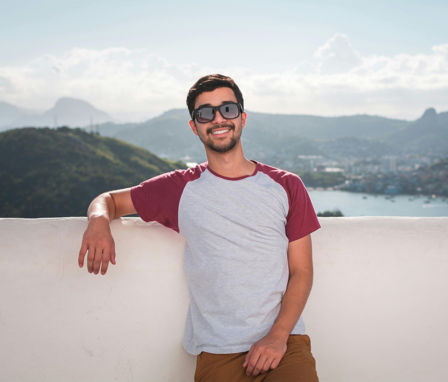 Man with sunglasses smiles, leans against a white wall, scenic background with mountains and water.