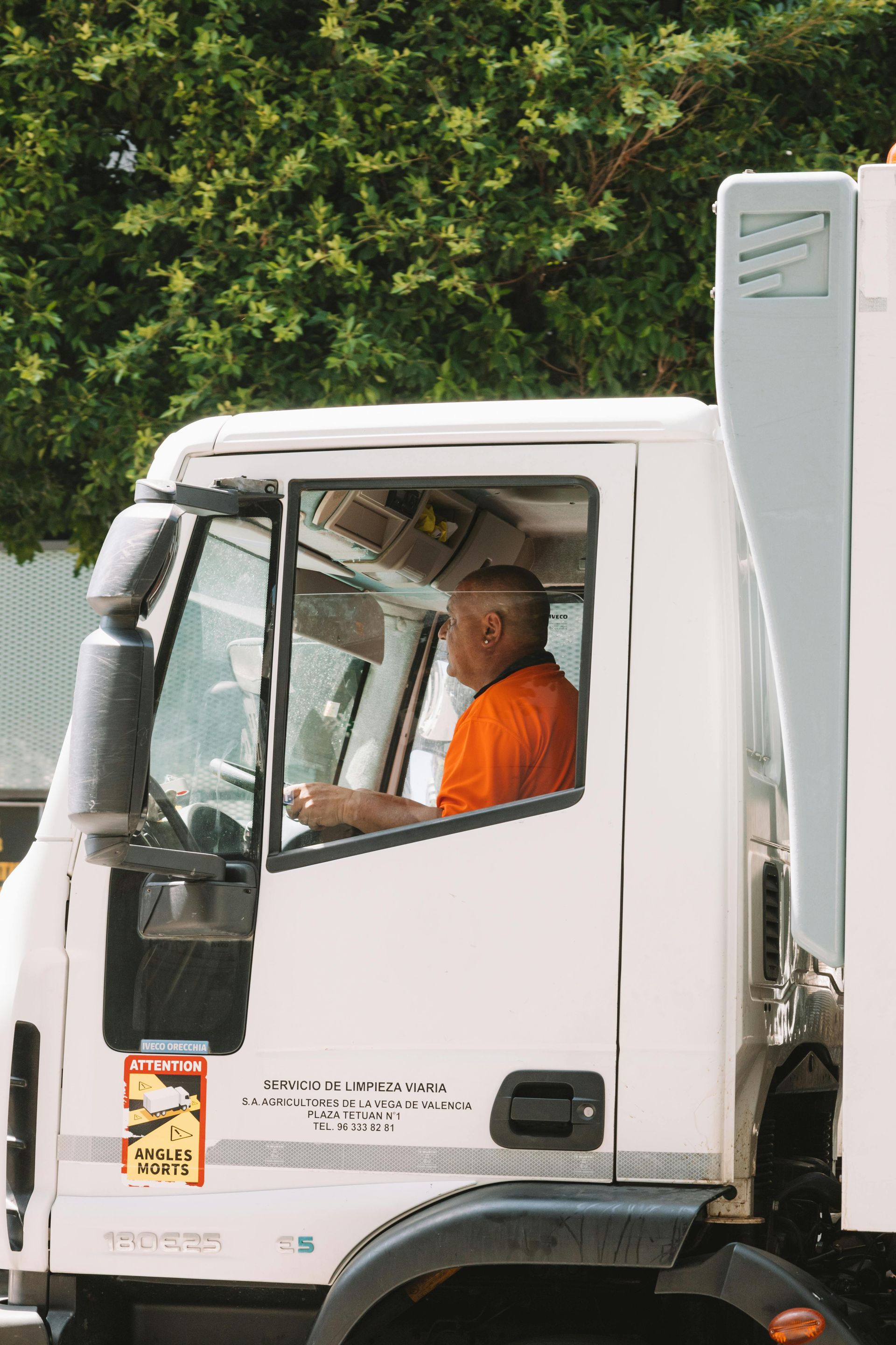 Man in orange shirt driving a white truck with a tall side, tree background.